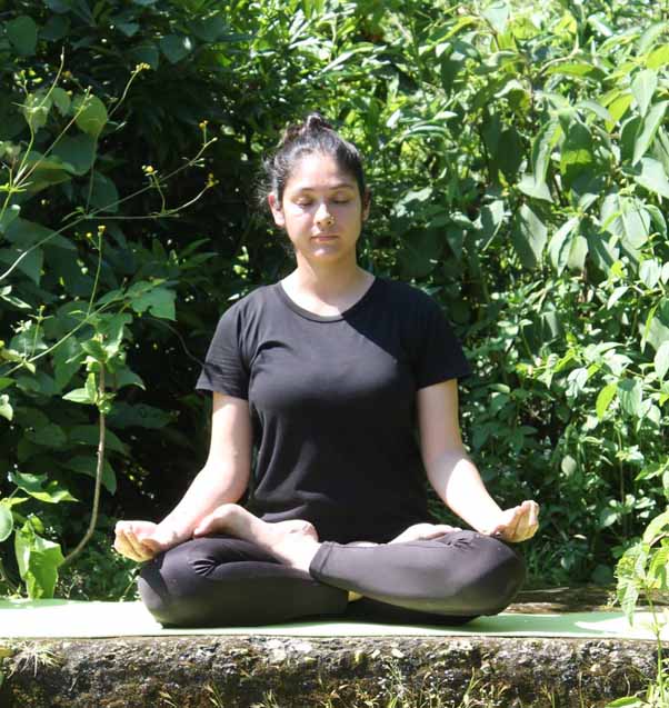 Acharya Indu in a seated yoga pose on a mat outdoors, demonstrating meditation with a straight back and hands in mudra, surrounded by nature in soft natural light.