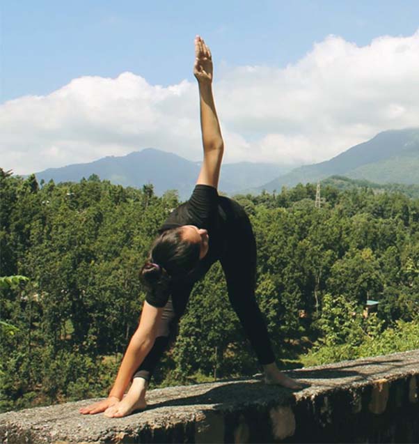 Acharya Indu performing a Triangle Pose (Trikonasana) outdoors on a ledge with lush green forest and mountain scenery in the background, under a clear sky, demonstrating balance and flexibility in nature.